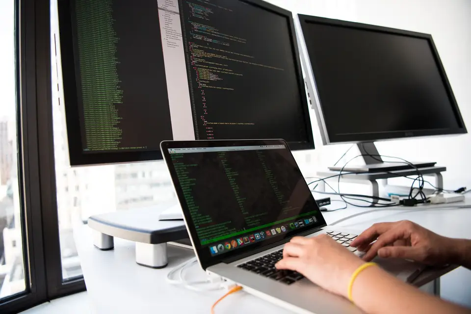 Entwickler schreibt Code am Laptop vor zwei großen Monitoren